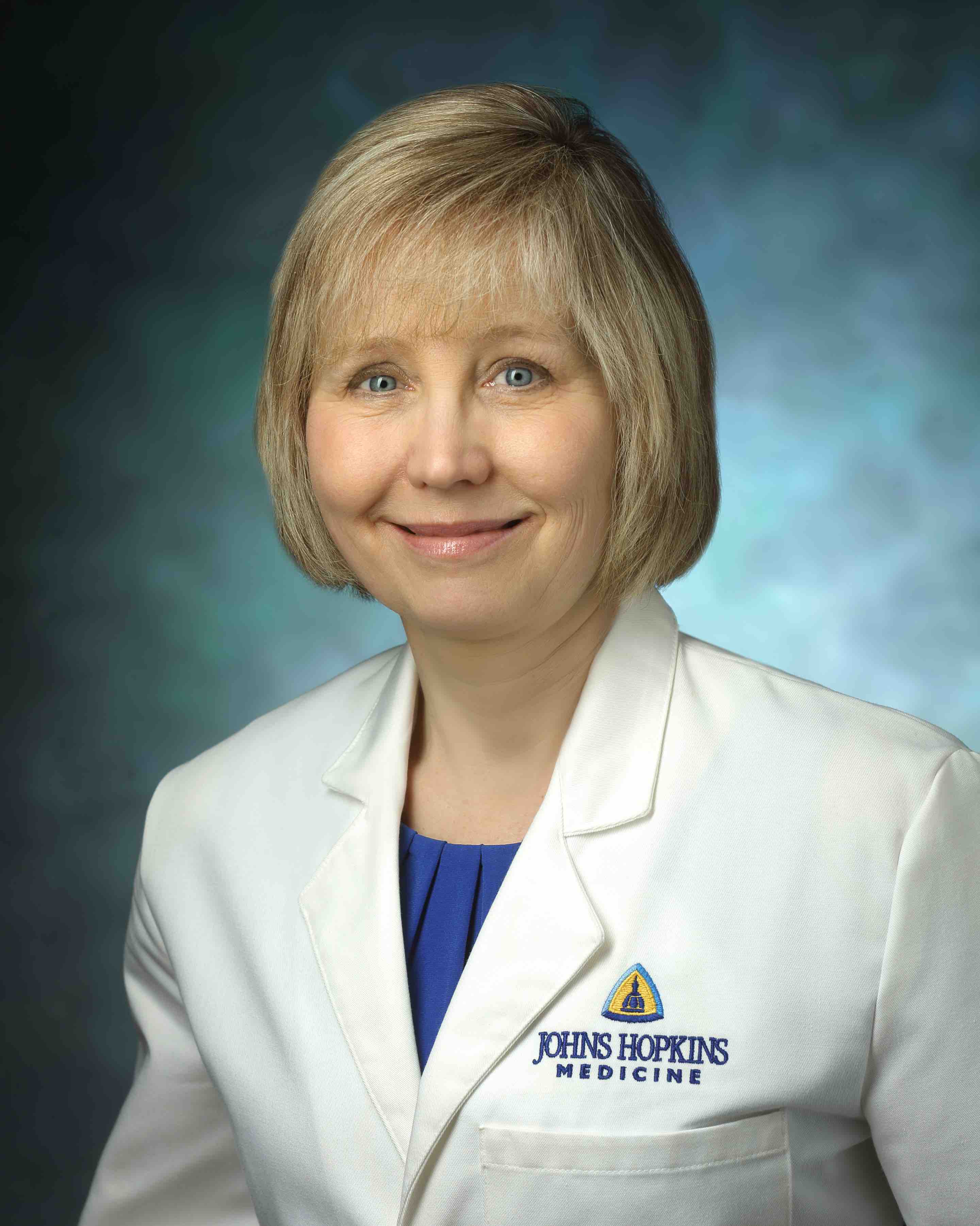 Dr. Valerie Baker wearing a traditional white lab coat with a blue blouse underneath on a blue background.