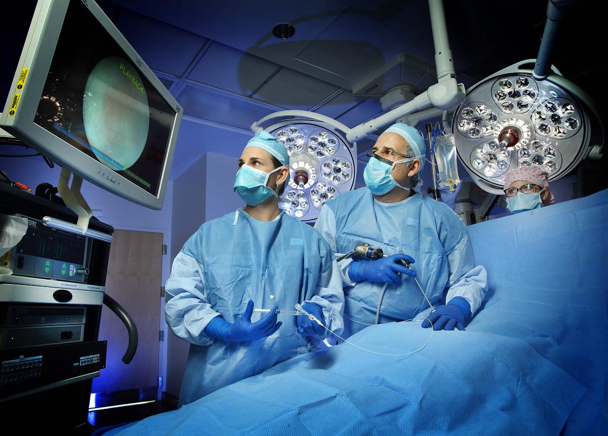 Jena Miller and Ahmet Baschat demonstrate a fetoscope in an OR setting. Both are wearing surgical gowns, caps, masks and gloves and are looking at a monitor screen.