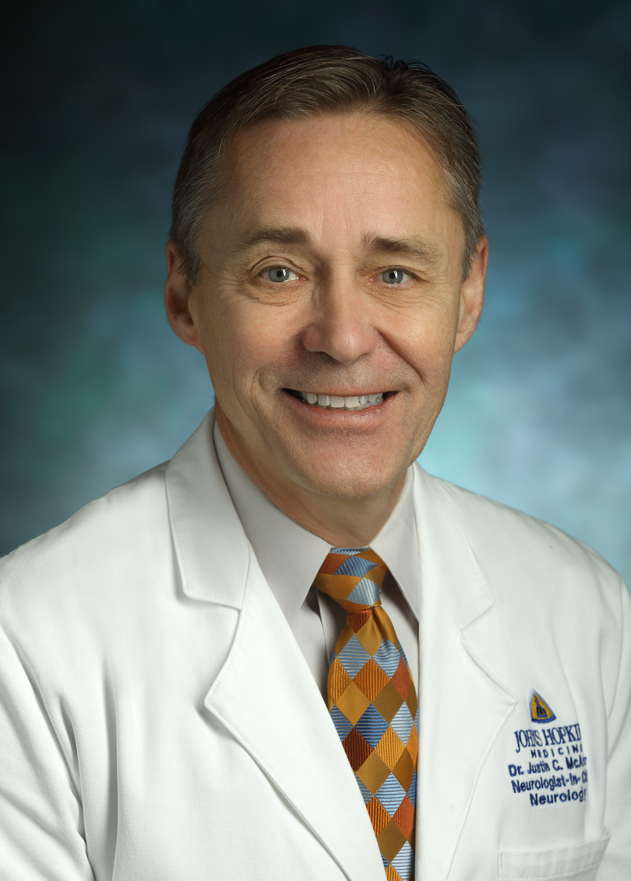 Justin McArthur in a formal portrait wearing a white lab coat, light gray button down shirt and multi-color checkered tie